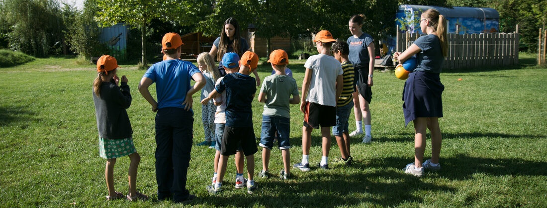 mehrere Kinder stehen auf einer Wiese mit 3 Erwachsenen; sie erhalten eine Einführung für ein Ballspiel