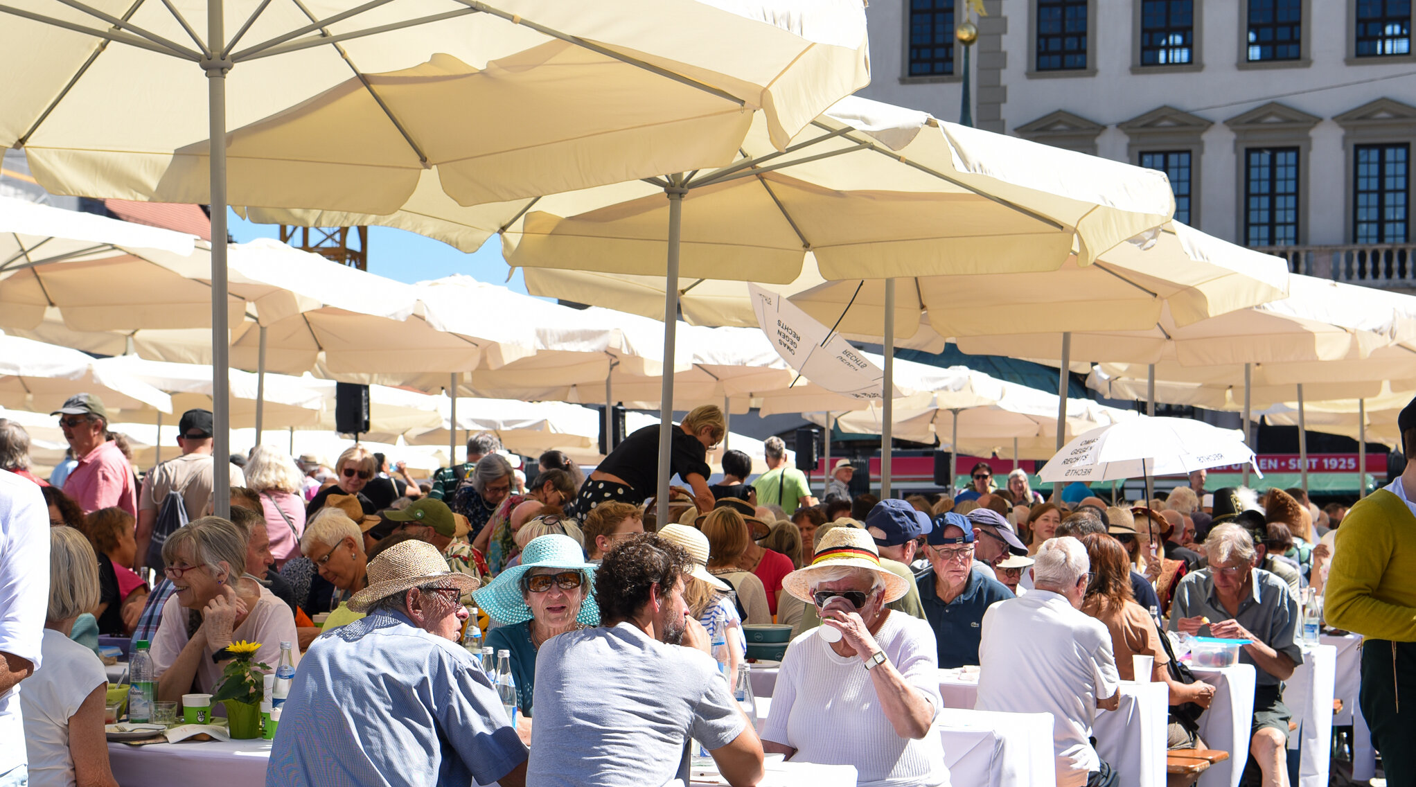 viele Menschen sitzen zusammen unter Sonnenschirmen an Biertischen, im Hintergrund ist das Augsburger Rathaus zu erkennen