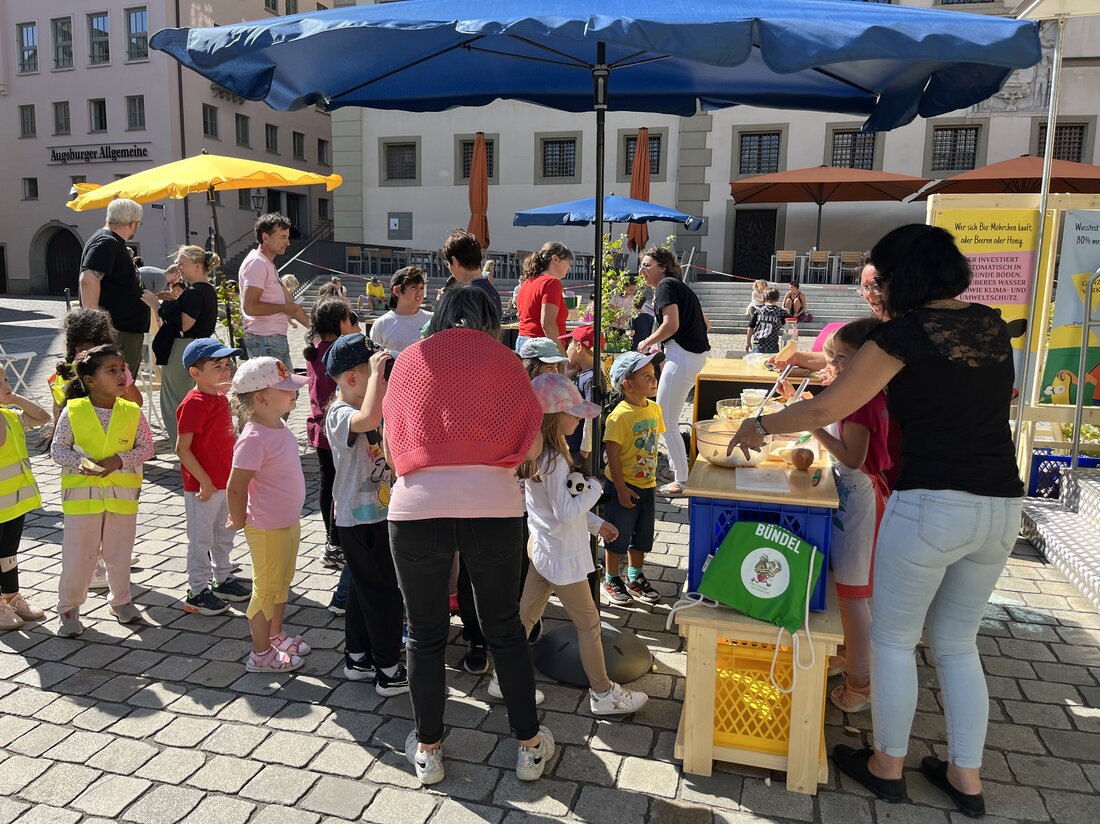 Auf dem Elias-Holl-Platz schauen Kinder im Grundschulalter zu, wie Erwachsene Lebensmittel zubereiten.