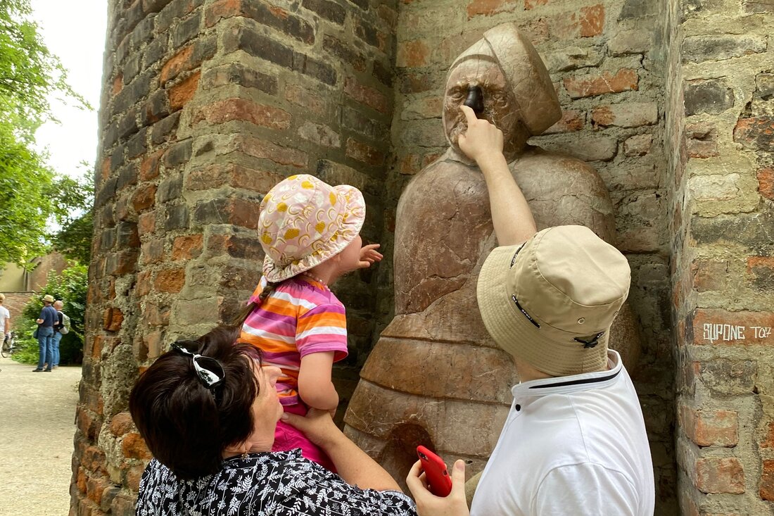 Eine Familie steht vor der Stadttue des "steinernen Manns" an der Augsburger Stadtmauer. Die Mutter links hebt ein Mädchen hoch zum Gesicht der Statue. Der Vater rechts berührt mit der rechten Hand das Gesicht der Statue. 
