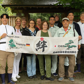 Eine Gruppe von Europäern und Asiaten hält gemeinsam ein Banner, auf dem Augsburg und Amagaski steht