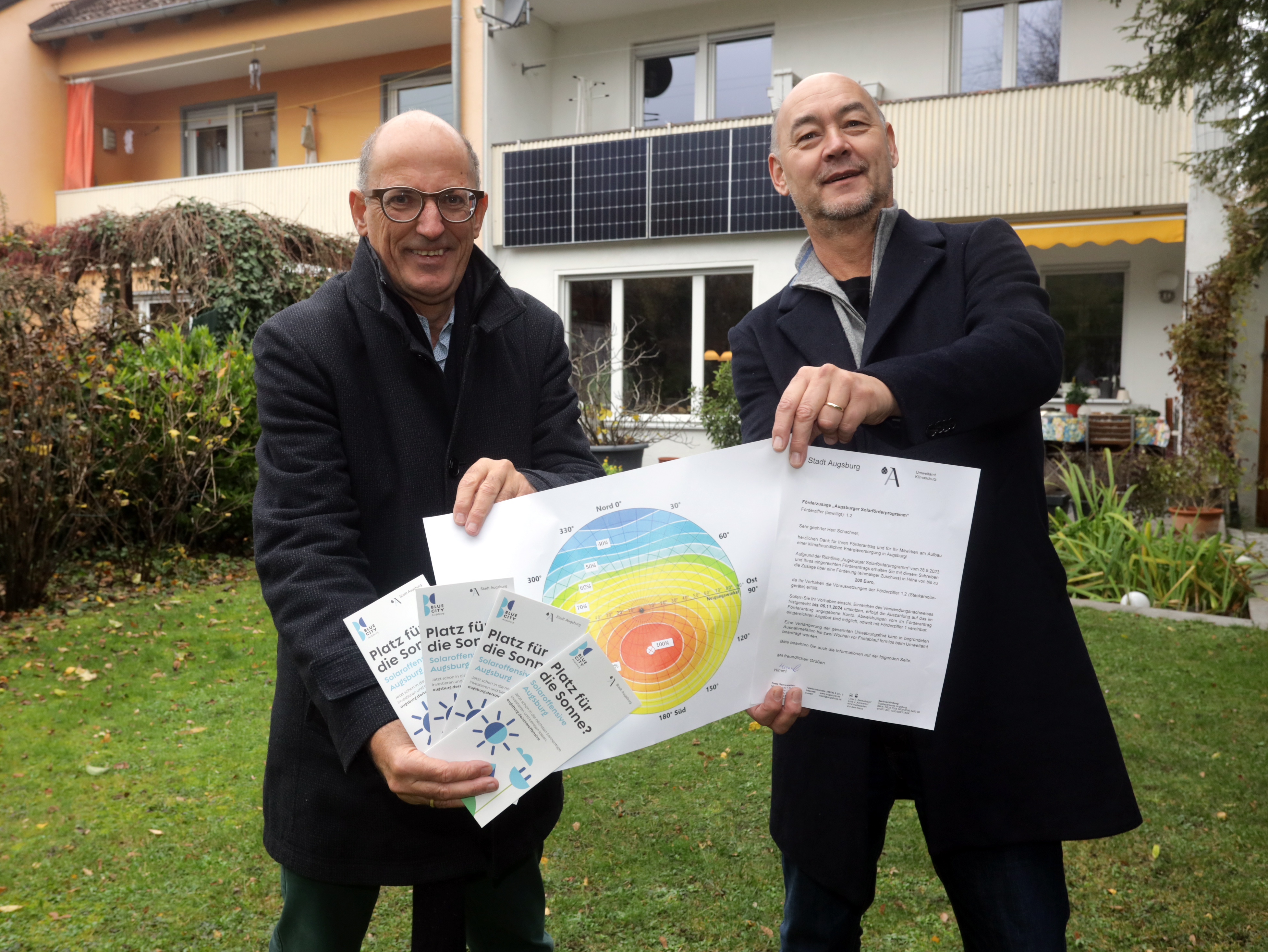 Zwei Männer vor einem Haus. Am Balkon ist eine Solaranlage zu sehen. Die Männer halten Unterlagen in der Hand.