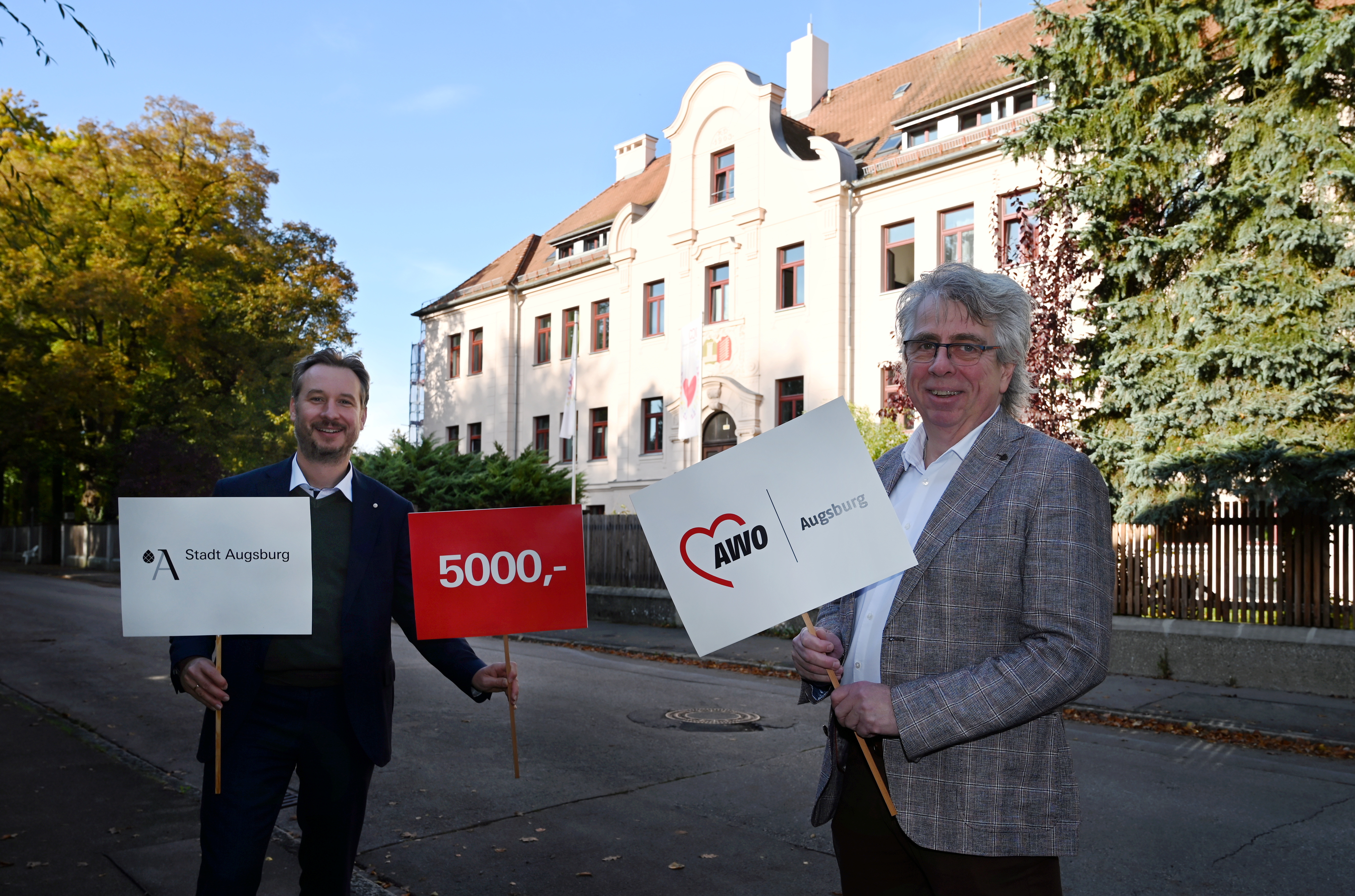 Zwei Männer vor einem Historischen Haus bei schönem Wetter. Sie halten Schilder mit den Aufschriften Stadt Augsburg 5000 und AWO in der Hand.