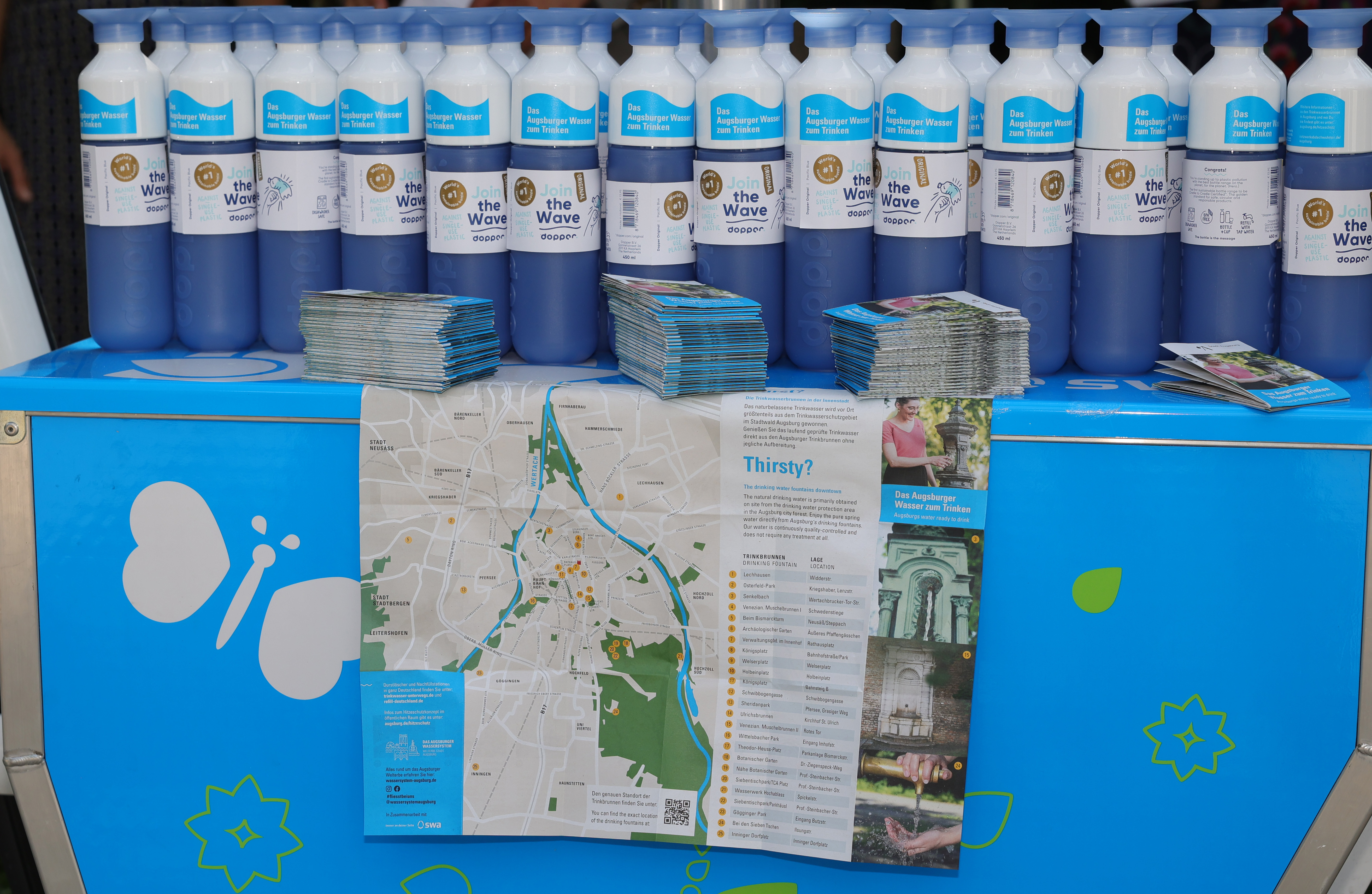 eine Reihe Trinkwasserflaschen vor einem Faltplan, der die Standorte der Trinkwasserbrunnen in Augsburg auflistet