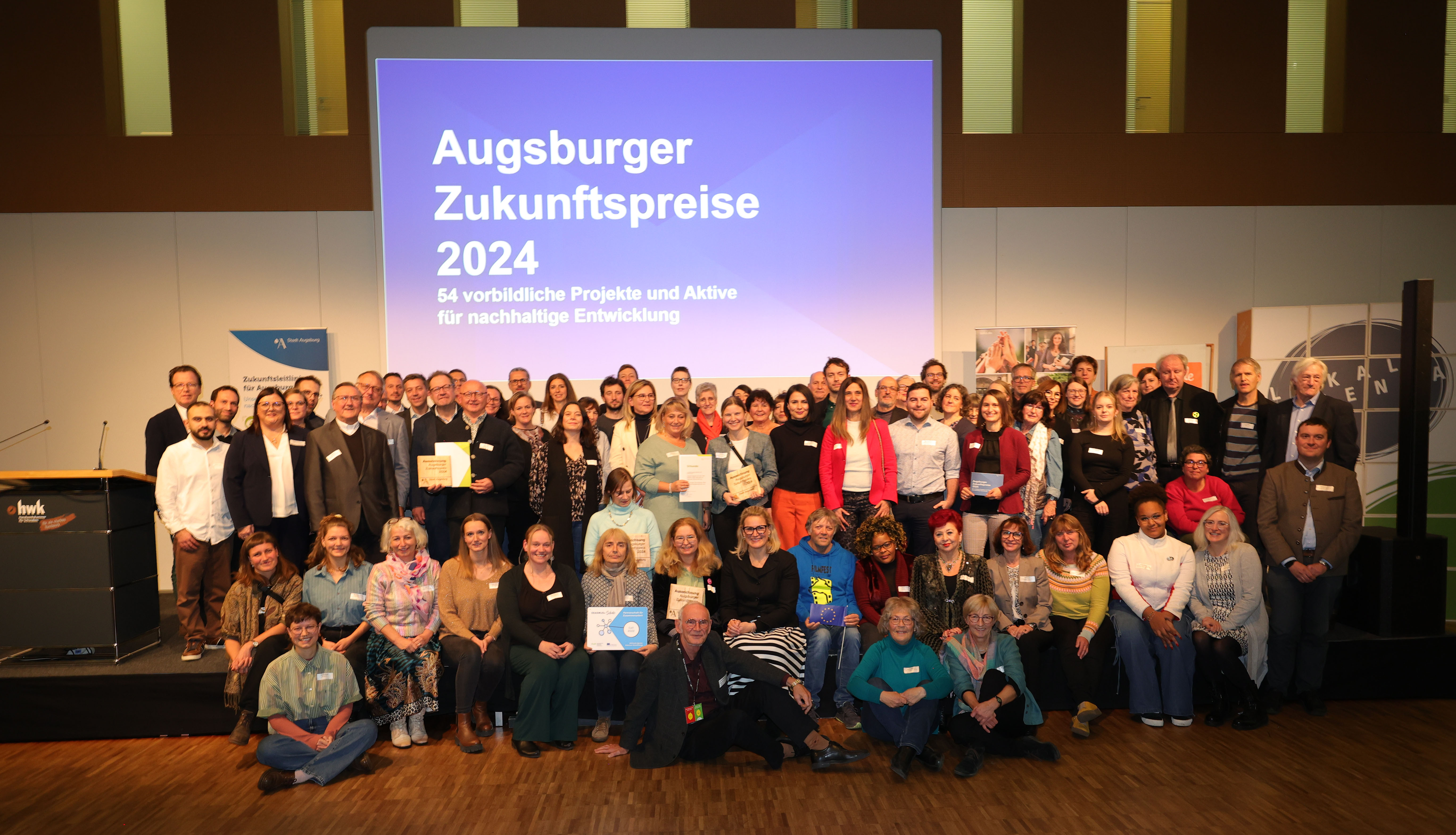 Etwa 70 Menschen vor einem großen Bildschirm. Auf diesem Steht "Augsburger Zukunftspreise 2024. 54 vorbildliche Projekte und Aktive für nachhaltige Entwicklung