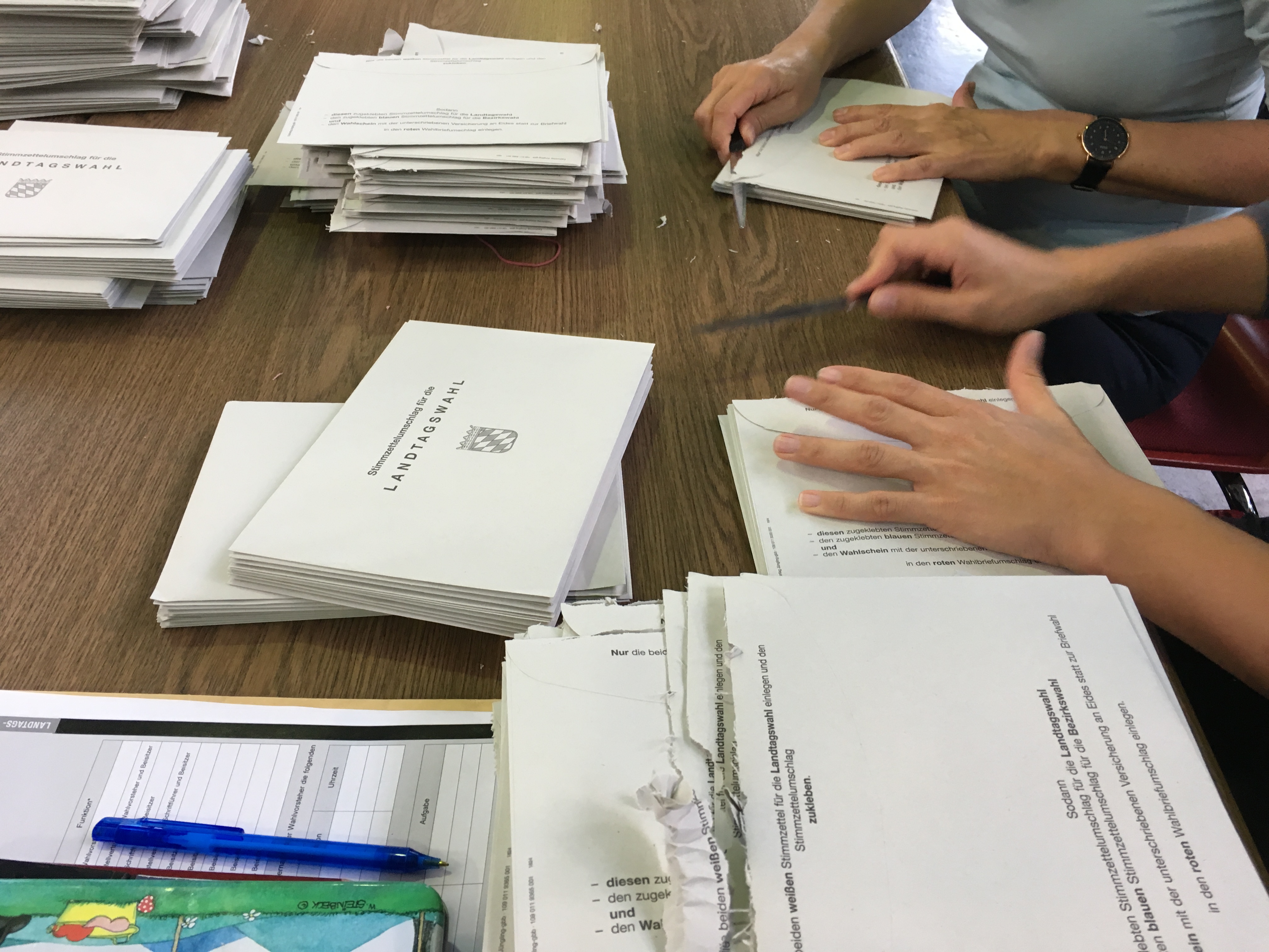 Auf einem Tisch liegen zahlreiche Briefumschläge mit der Aufschrift "Landtagswahl". Rechts sind die Hände von zwei Personen zu sehen. Sie öffnen je einen Umschlag.