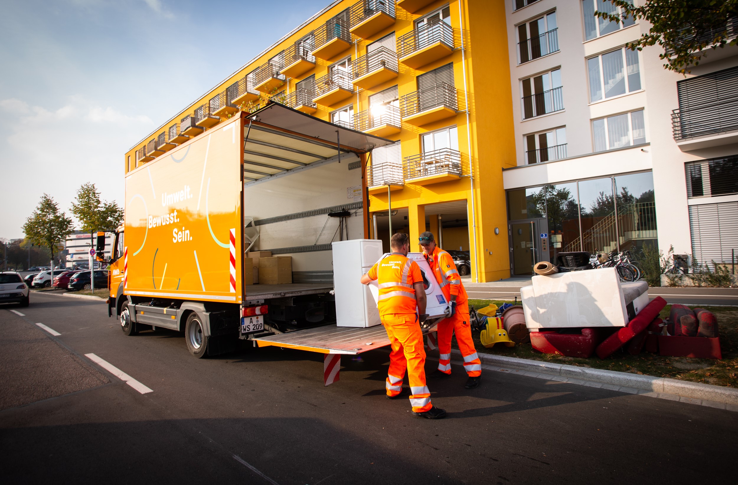 Zwei Männer in orangefarbener Kleidung heben ein Möbelstück in einen Lastwagen.