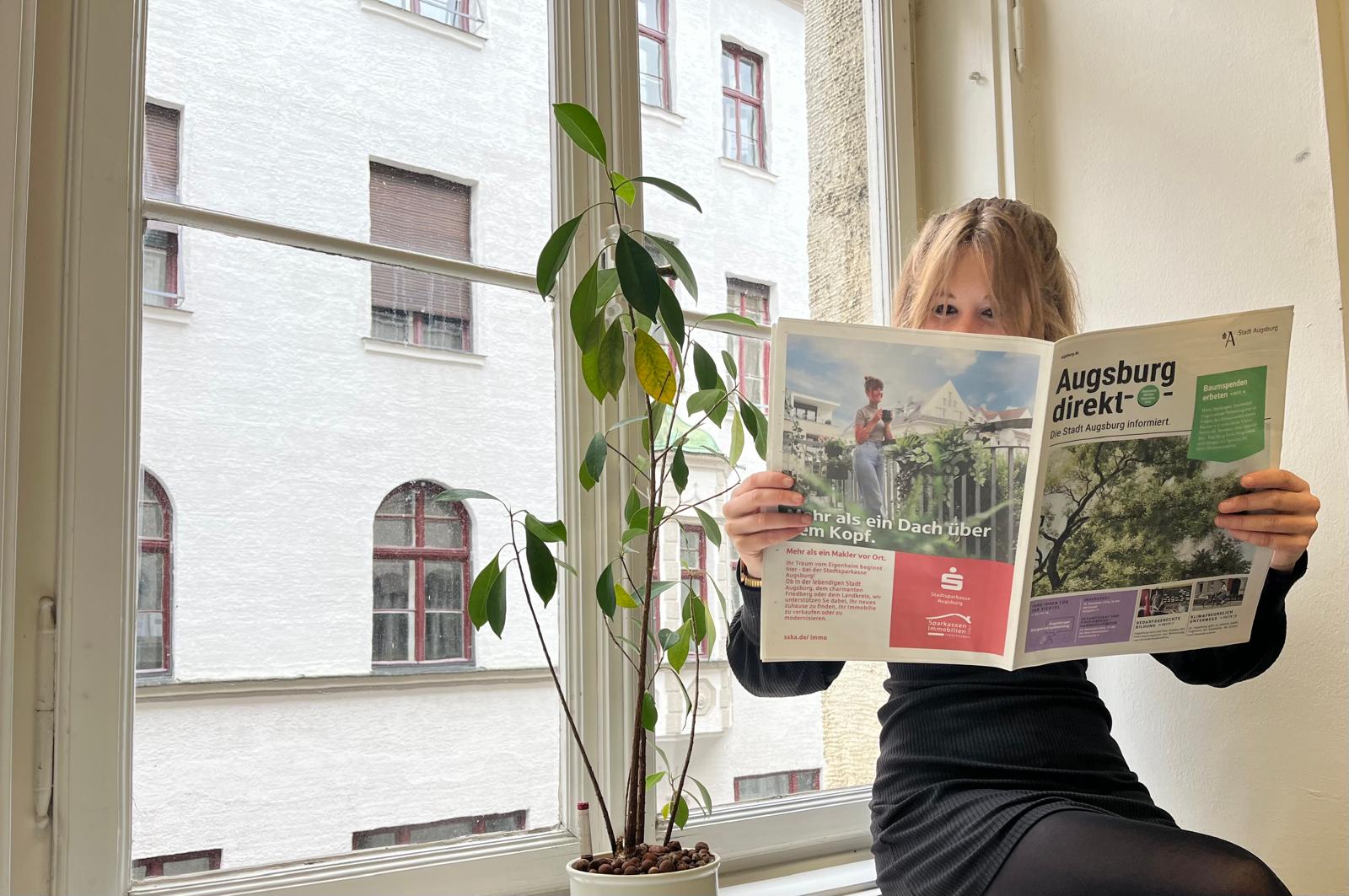 Eine frau sitzt auf einer Fensterbank und liest die neue Augsburg direkt.