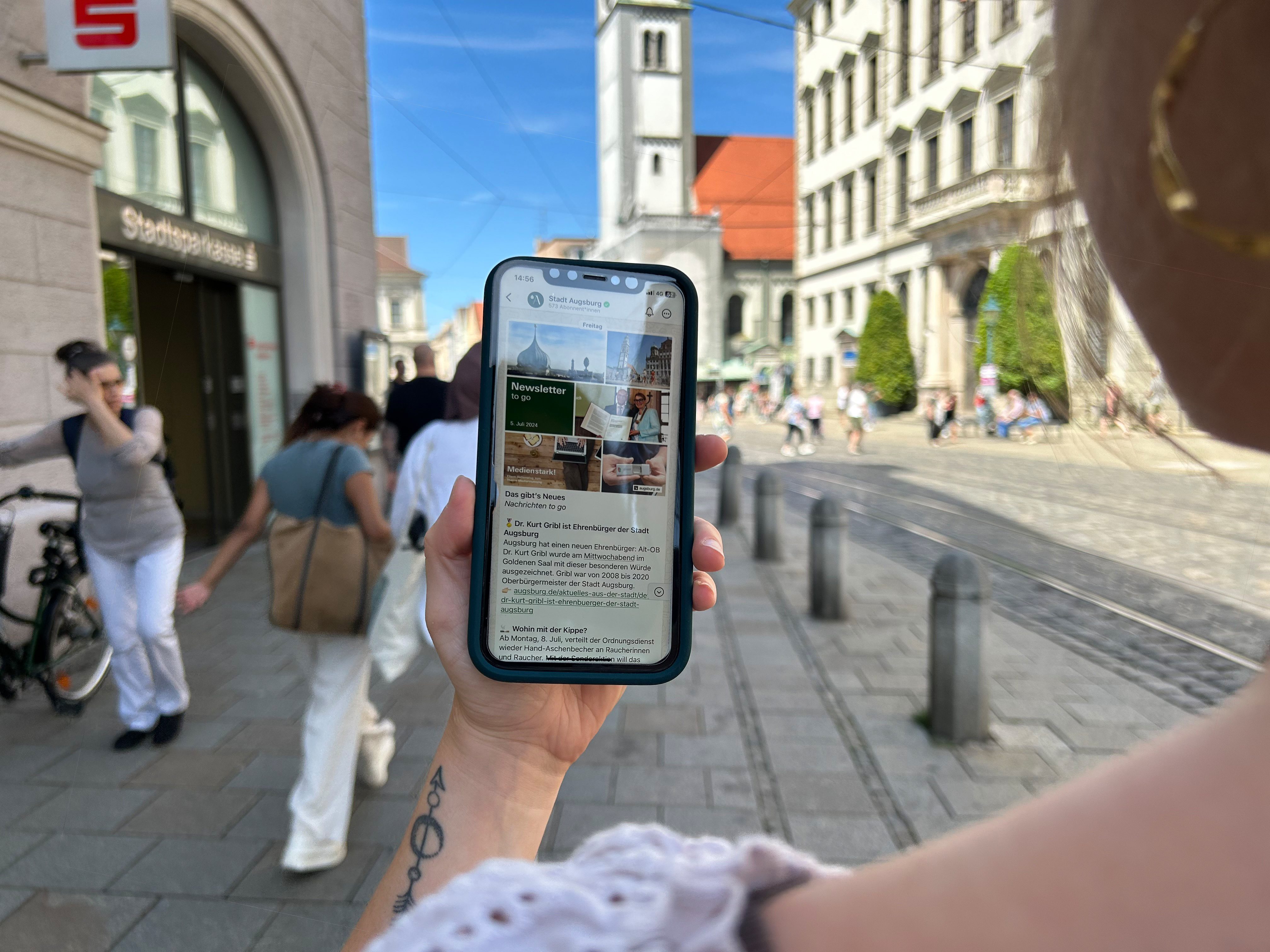 Ein Smartphone im Vordergrund, im Hintergrund das Augsburger Rathaus. Auf dem Display des Smartphones ist der WhatsApp Kanal der Stadt Augsburg zu sehen. 