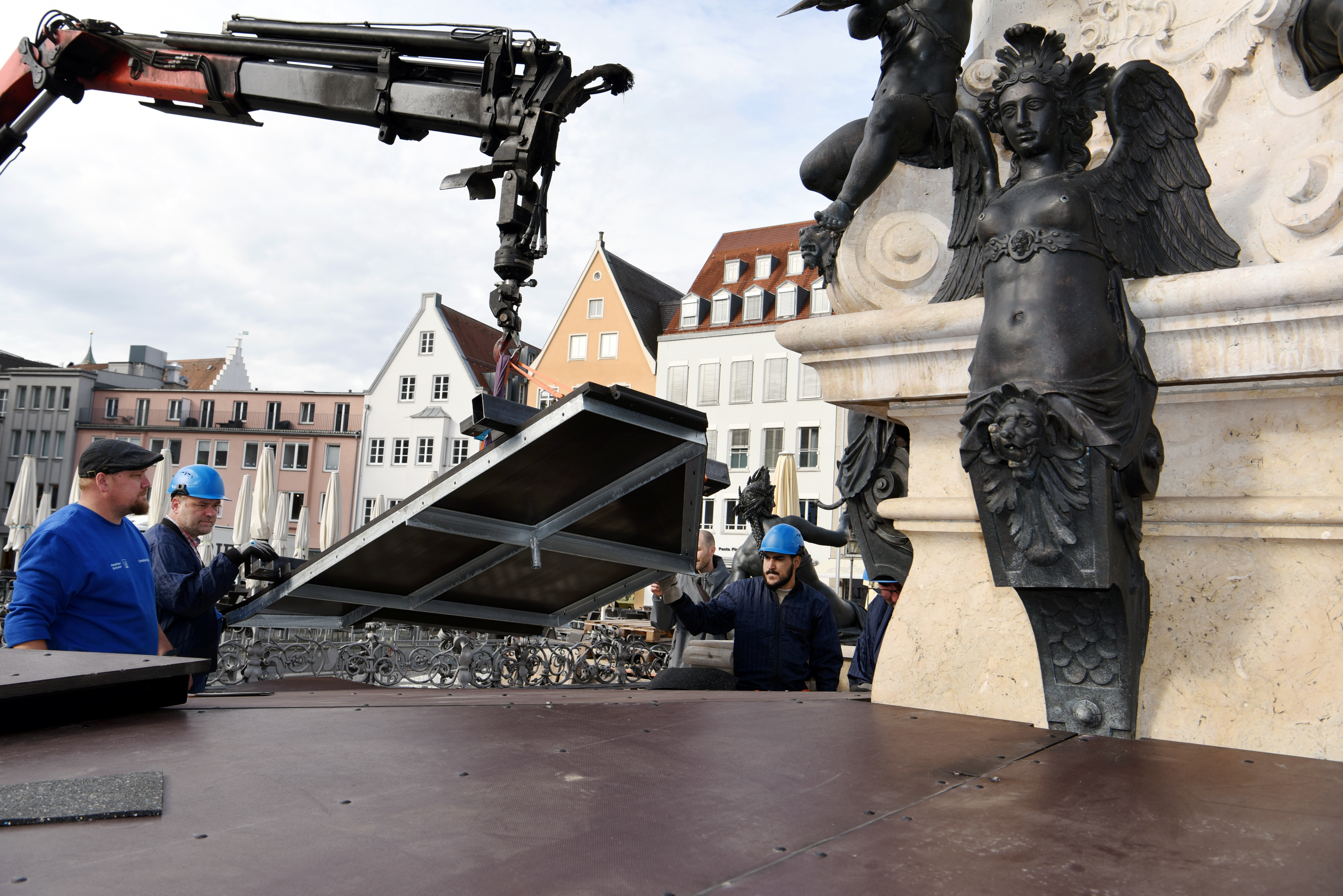 Männer in Arbeitskleidung heben mit einem Kran Metallplatten auf den Augustusbrunnen