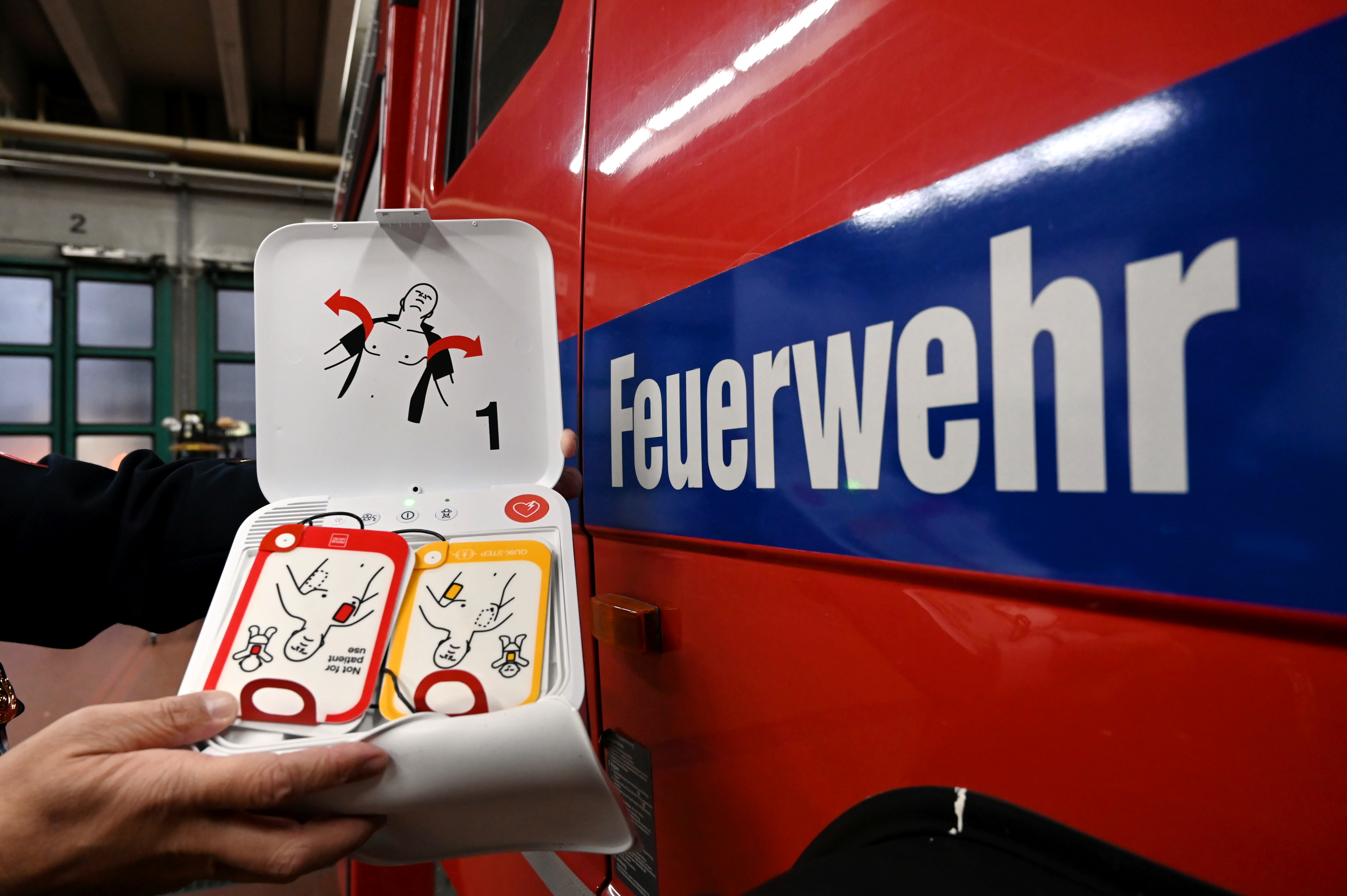 geöffneter Defibrillator neben einem roten Fahrzeug mit der Aufschrift "Feuerwehr"