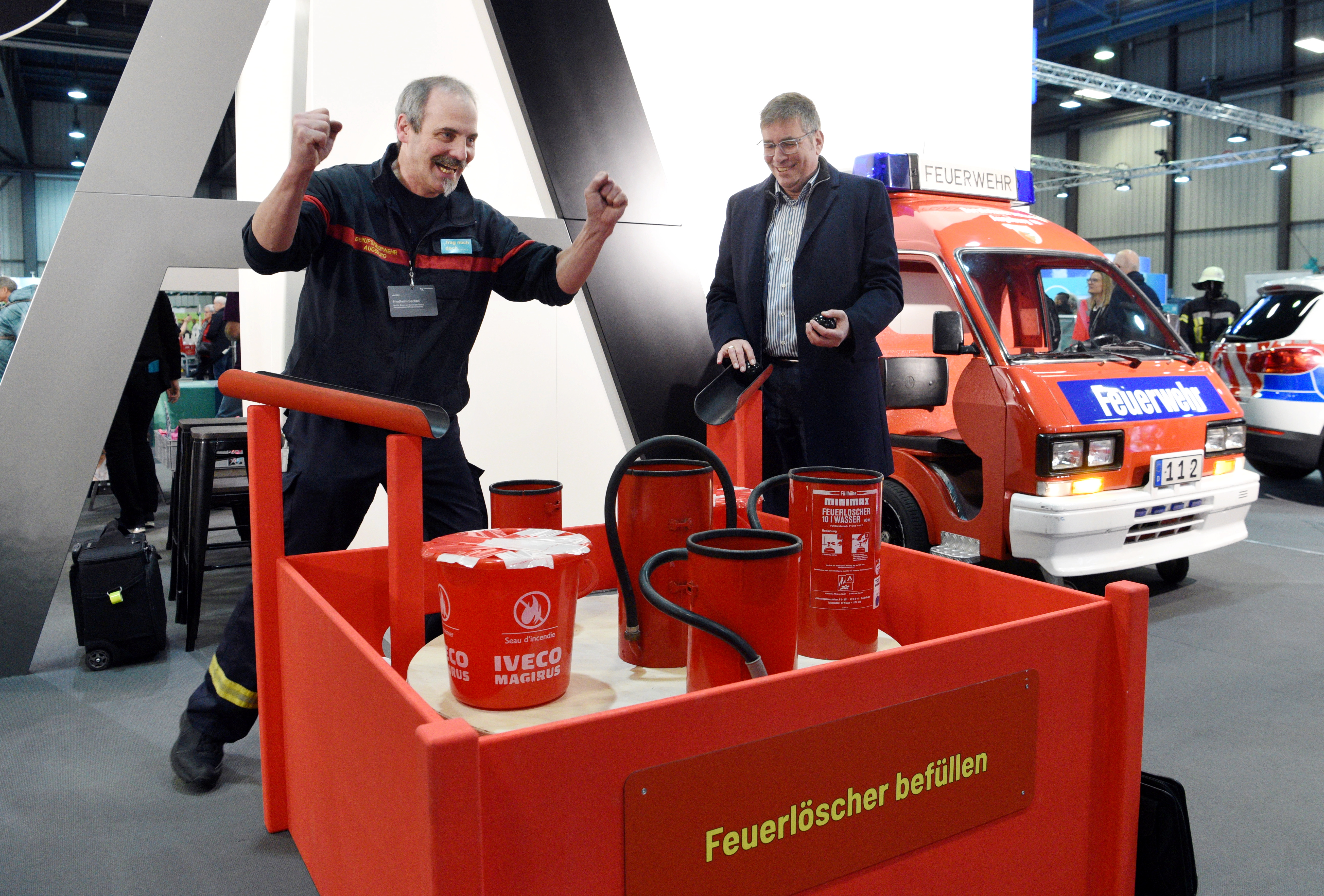 Ein Feuerwehrmann und ein Mann im Anzug stehen vor einer Roten Kiste mit mehreren Feuerlöschern.