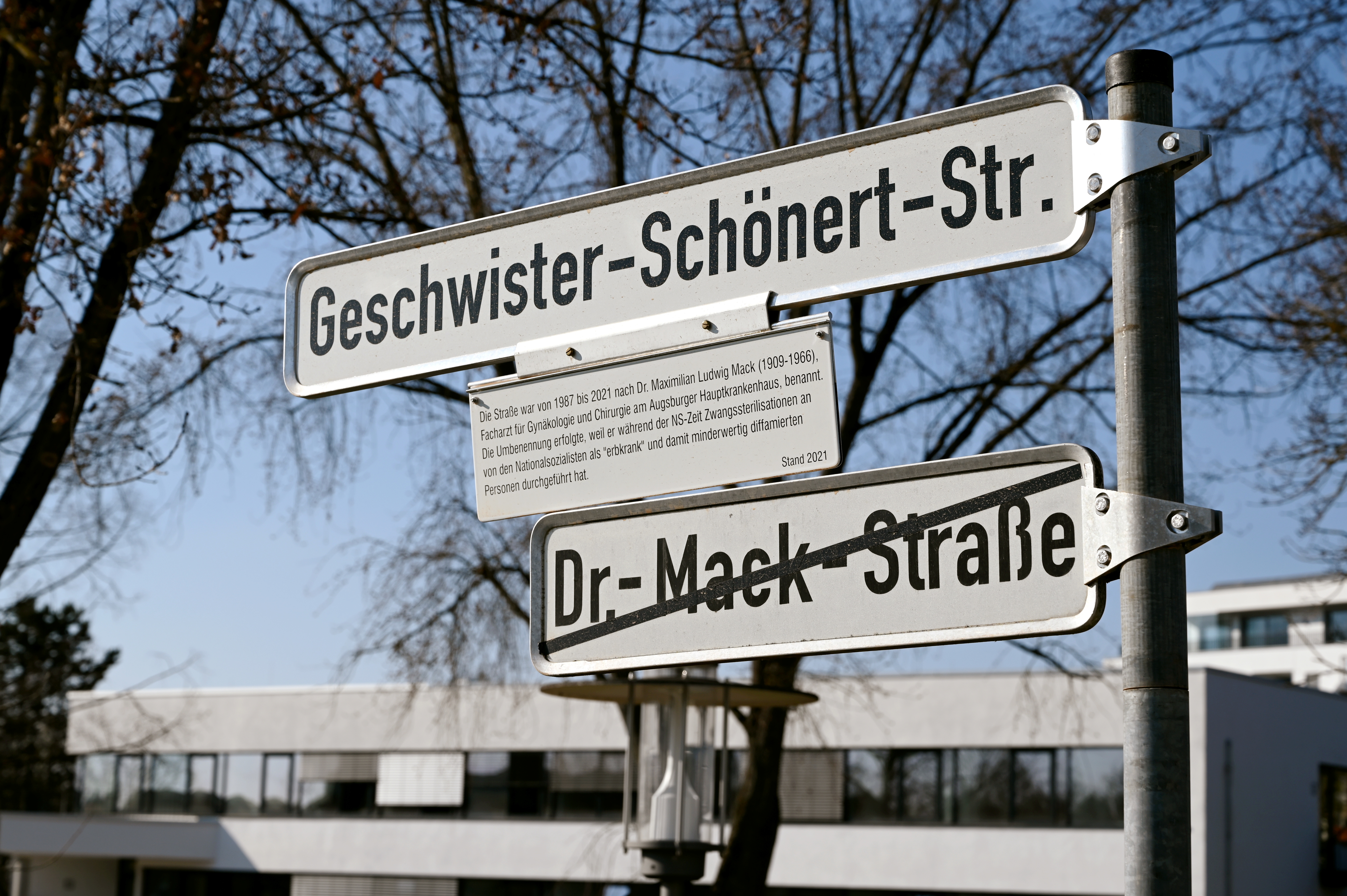 Zwei Straßennamen an einem Pfosten, der untere ist durchgestrichen, der obere durch einen Erklärtext ergänzt