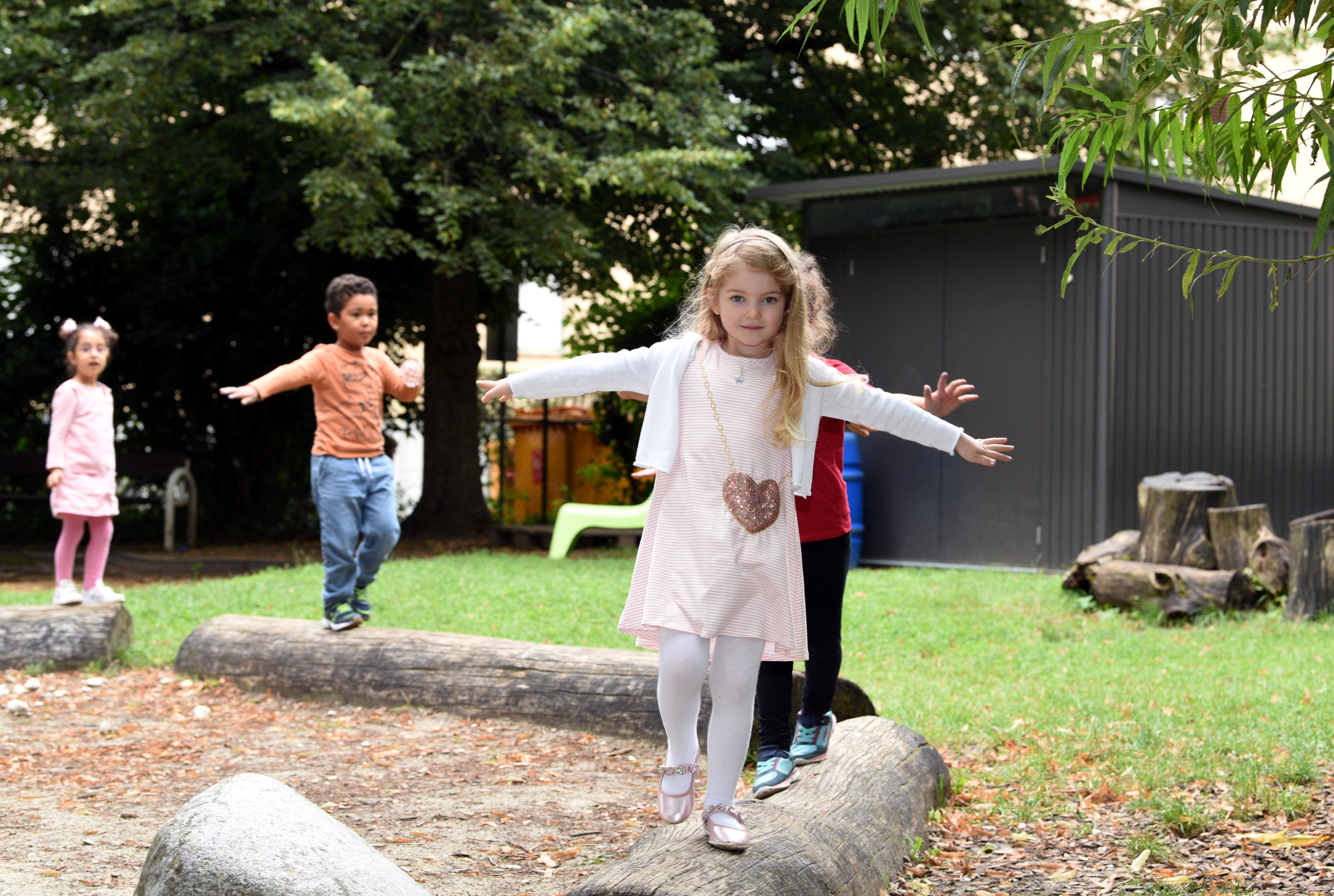 Vier Kinder balancieren über einen Baumstamm. Im Hintergrund ist ein Kindergarten zu sehen.