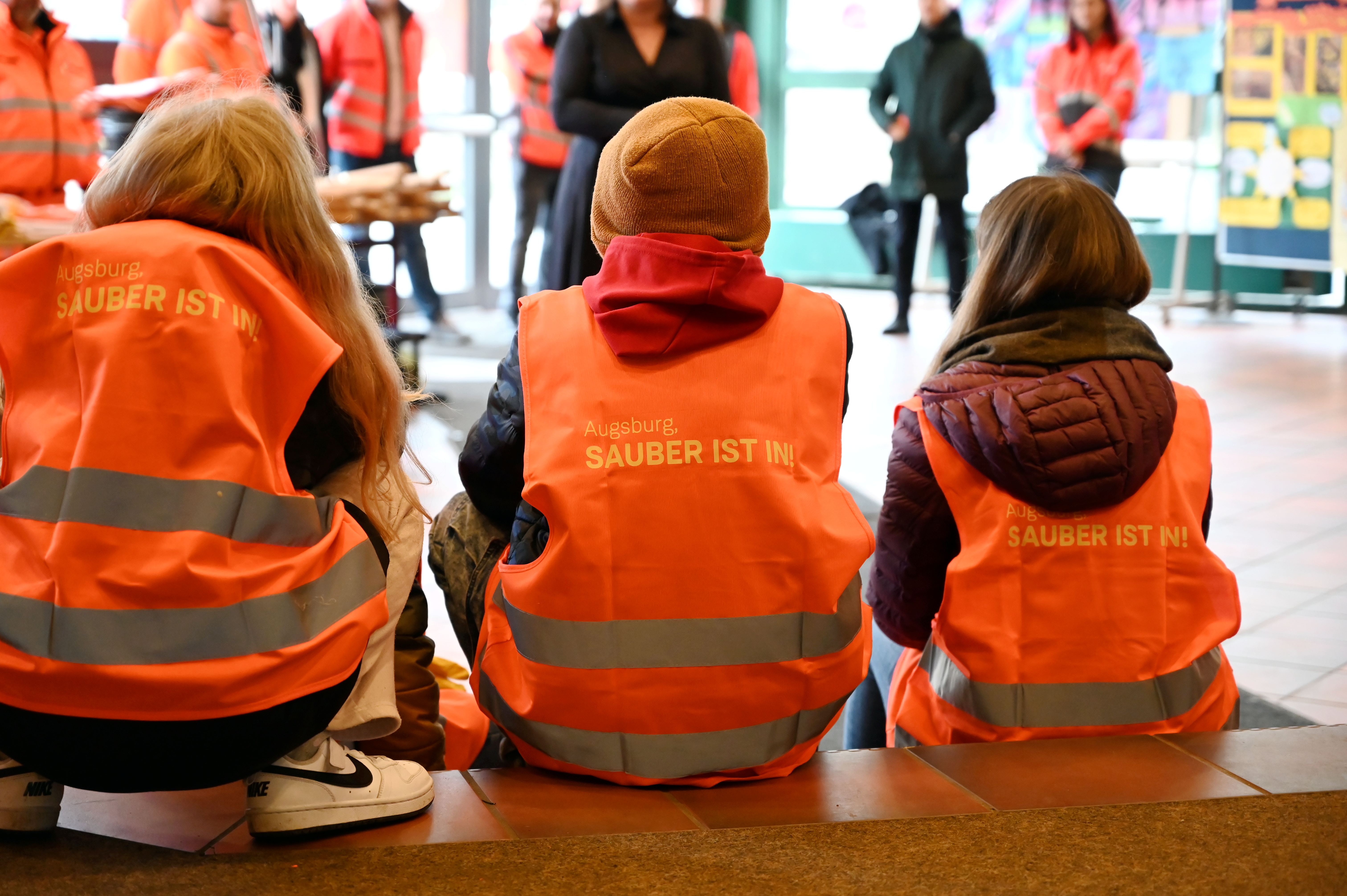 3 Kinder sitzen mit dem Rücken zum Betrachter nebeneinander; auf ihren Jacken steht "Sauber ist in"