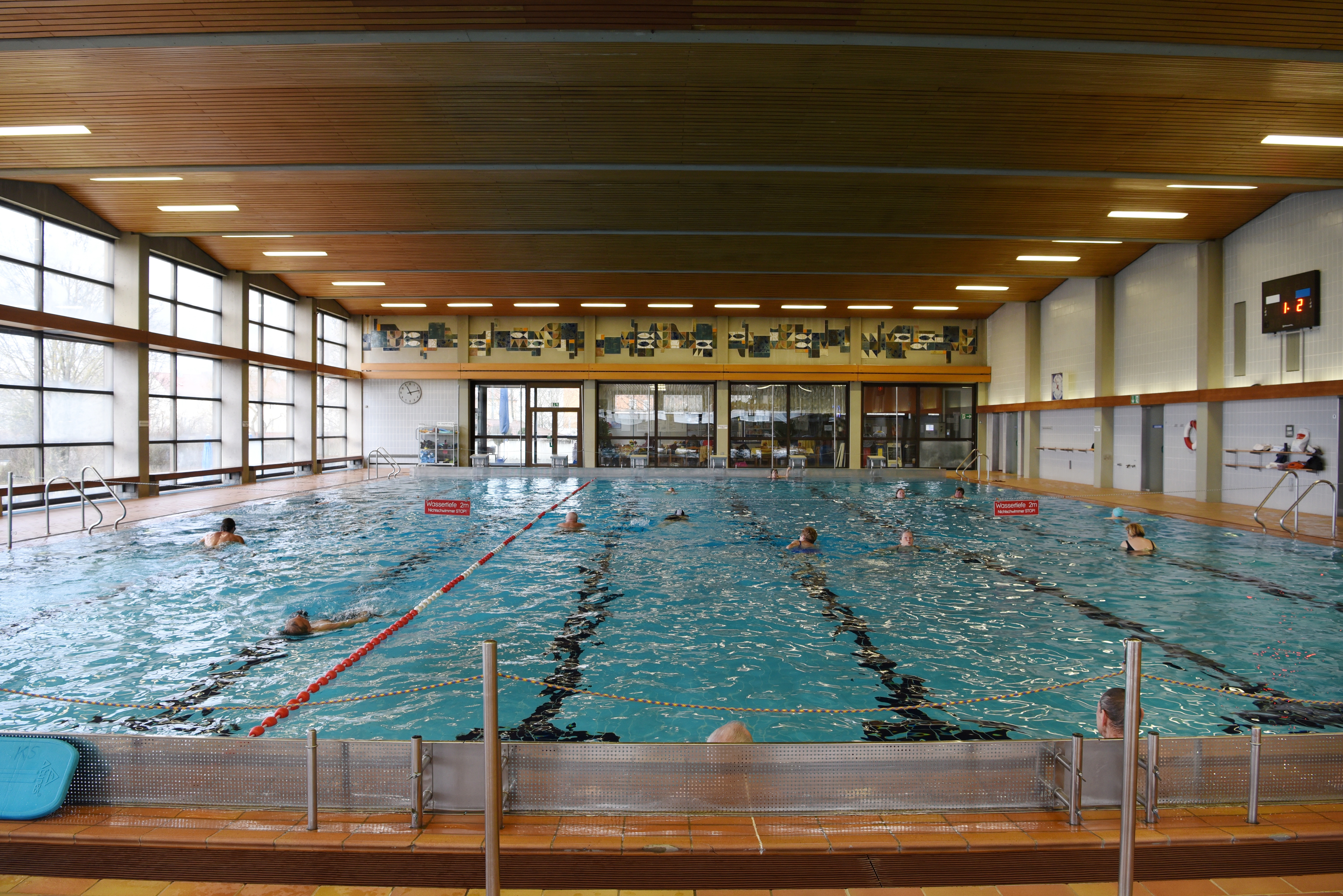 Es ist ein altes Hallenbad von innen zu sehen. Im Wasser schwimmen mehrere Menschen.