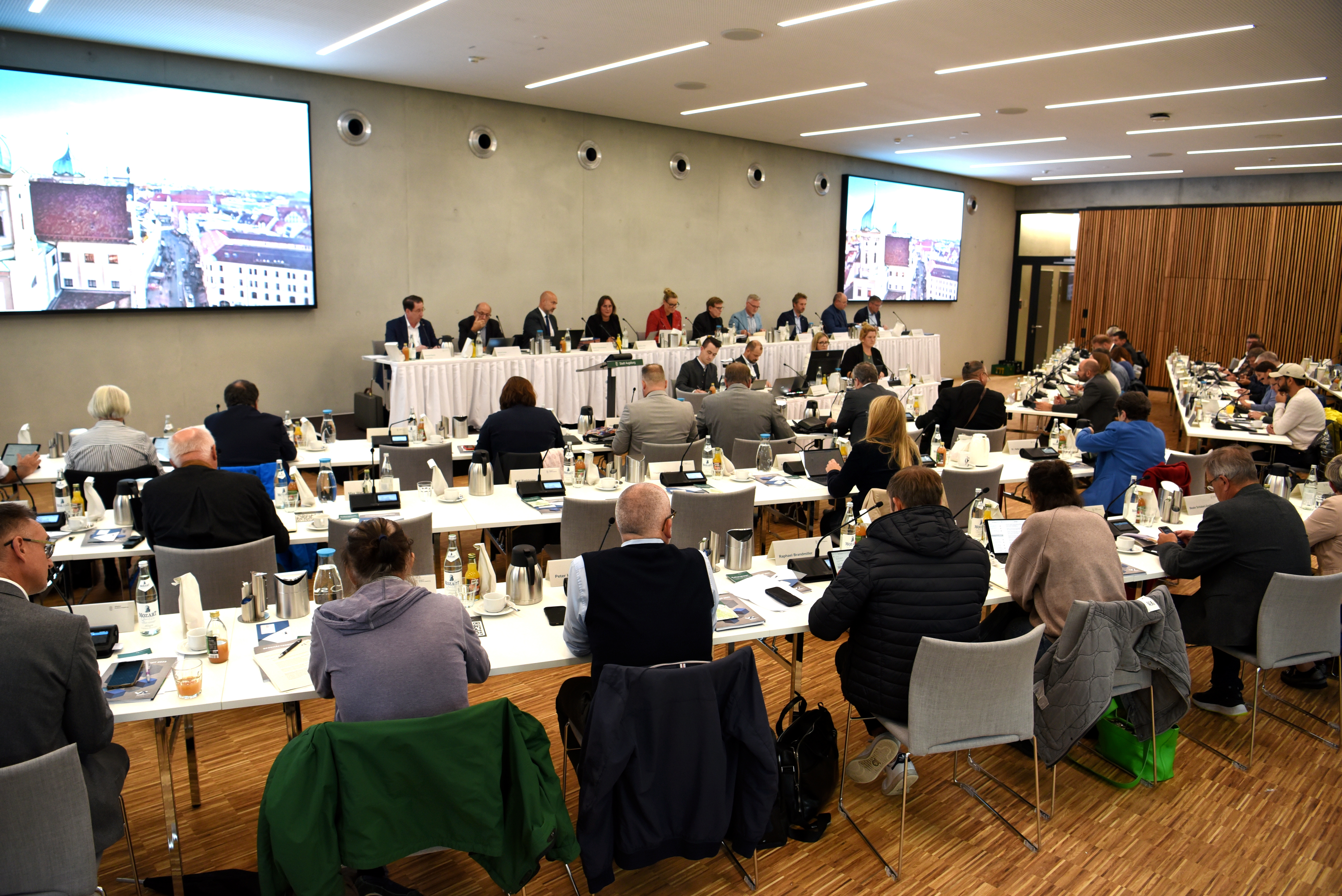 Viele Menschen sitzen in Tischreihen in einem Saal. An der Wand sind große Bildschirme zu sehen.