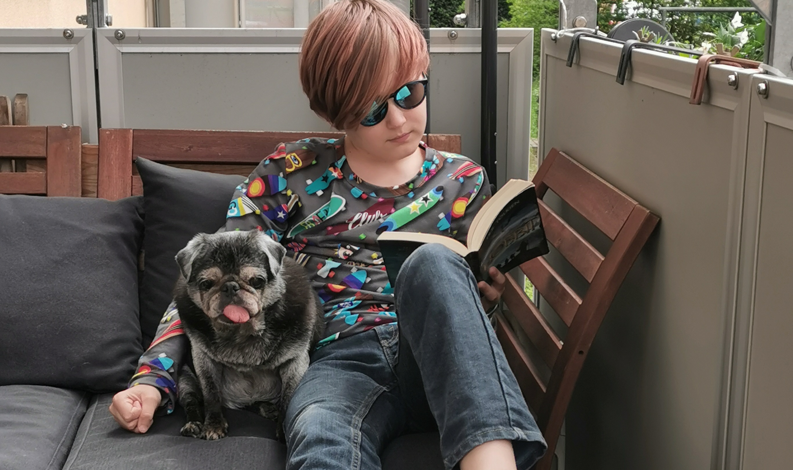 Ein Kind sitzt auf dem Balkon und liest ein Buch. Ein Hund sitzt neben dem Kind.