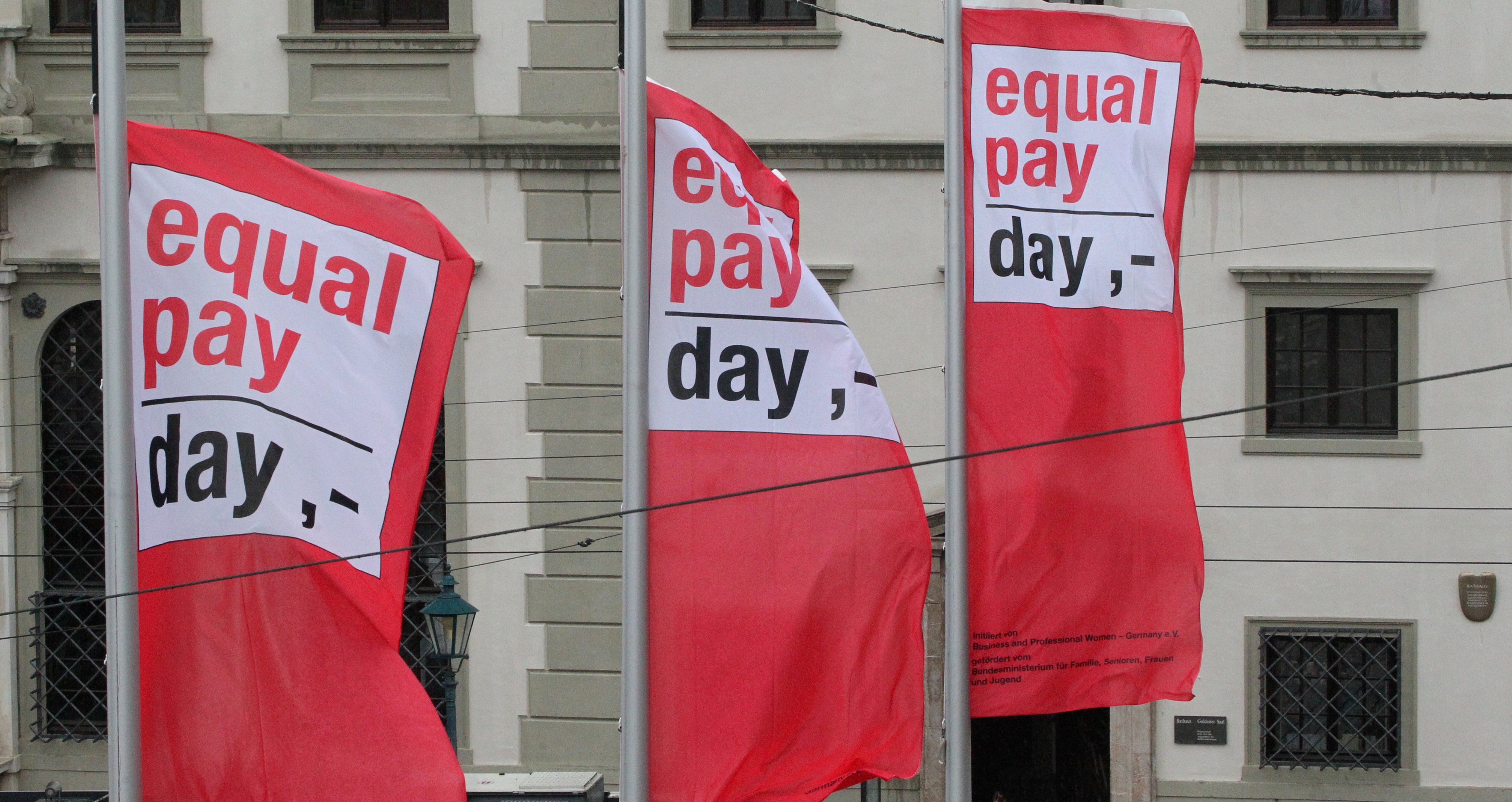 Drei Flaggen wehen vor dem Rathaus. Sie tragen die Aufschrift "Equal Pay Day"