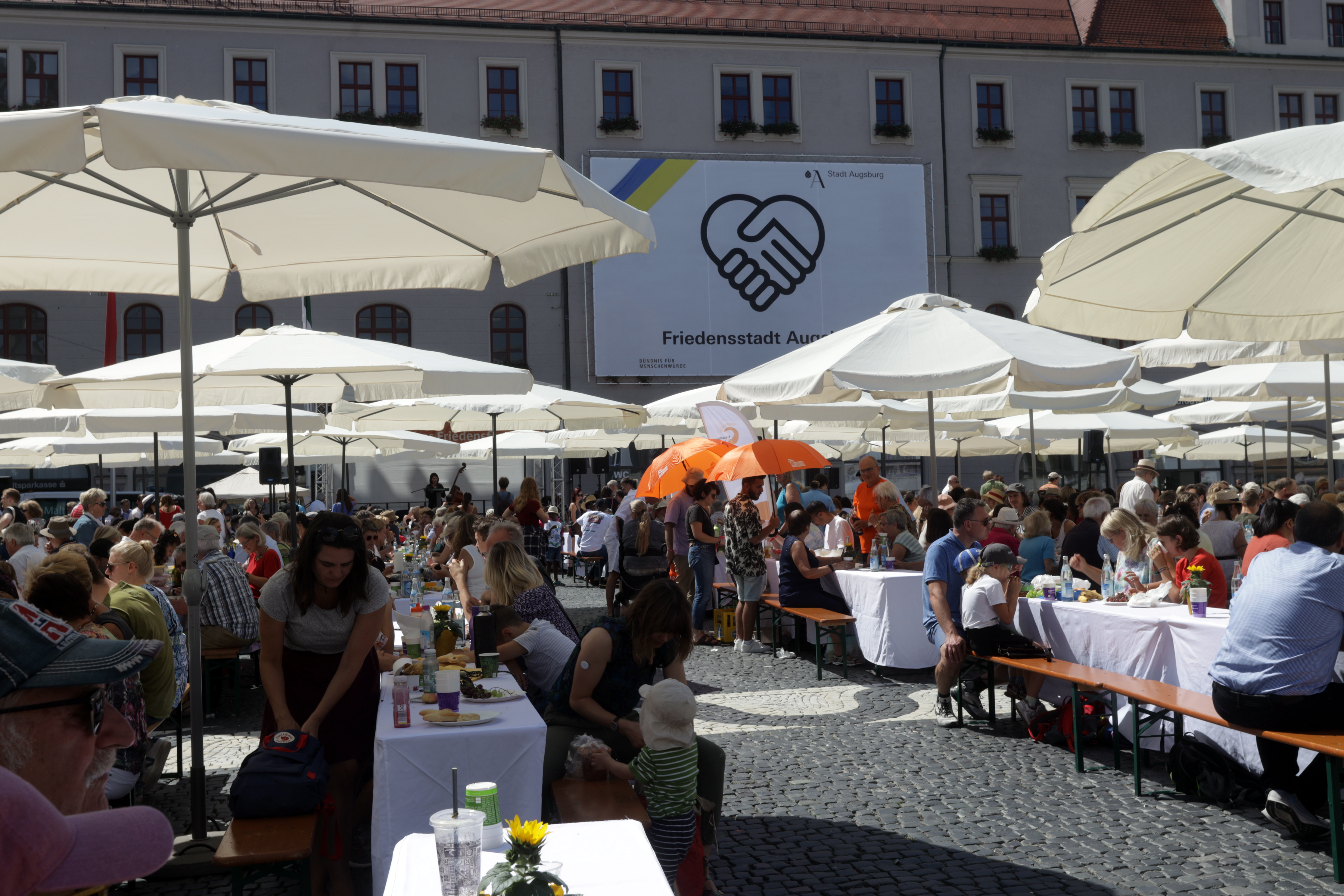 Aufgespannte Sonneschirm beschatten Bierbänke, an denen Menschen sitzen. Im Hintergrund ein Plakat "Friedensstadt Augsburg"